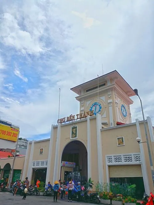 Hai bạn trẻ ngồi trên bậc thềm gần khu vực chợ đêm, vừa ăn vừa trò chuyện, phía sau là ánh đèn vàng hắt ra từ những quầy hàng