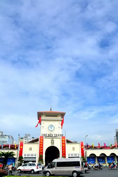 Khung cảnh mưa nhẹ trước cổng chợ, vài người trú dưới mái hiên, mặt đường phản chiếu ánh đèn tạo cảm giác rất khác so với ban ngày