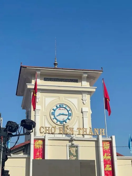 Một cô bán hàng đang gói đồ cho khách, tay làm nhanh nhưng vẫn nở nụ cười nhẹ, phía sau là những kệ hàng đầy ắp