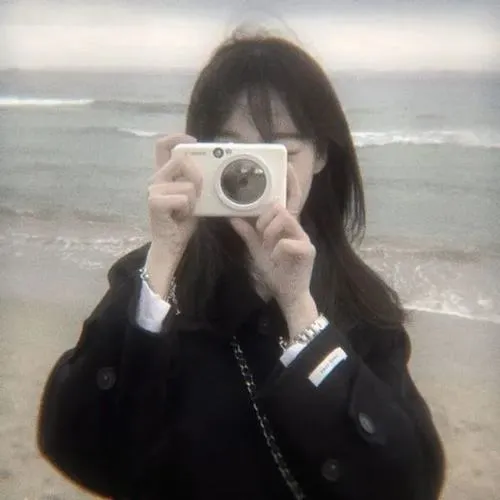 Gái xinh ôm gấu bông che miệng trong hoodie đen, tóc đen dài, tư thế ngồi co ro dễ thương.