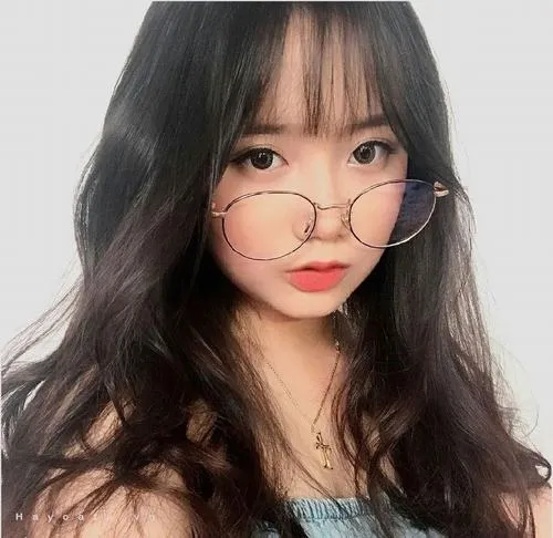 Gái xinh đeo kính gọng tròn mảnh ngồi tựa ghế trong phòng ánh sáng dịu, tay chống cằm nhẹ và nụ cười trong sáng trên gương mặt nhỏ nhắn.