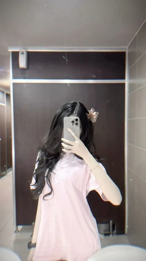 Tóc dài xõa mặc áo hồng nhạt đứng trong phòng sáng cầm điện thoại có hoa giấu mặt ấm áp.