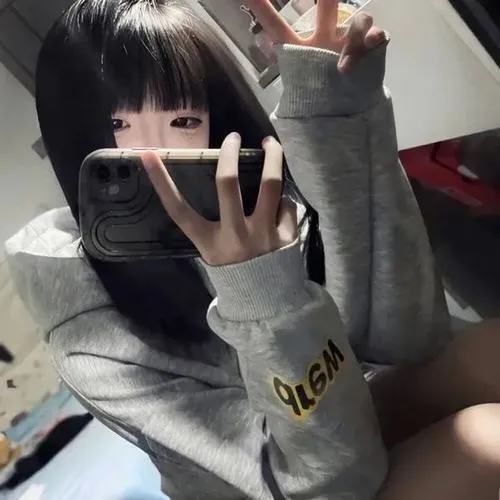 Ngồi tự nhiên tóc mái ngang áo xám oversized hai tay nâng điện thoại giấu mặt tạo cảm giác cá tính nhẹ nhàng.