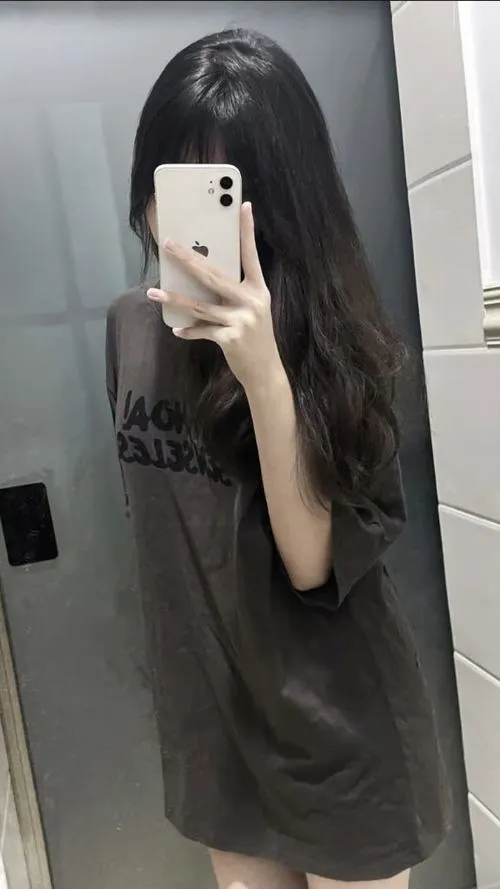 Tóc dài xõa mặc áo nâu rộng rãi dùng điện thoại che kín mặt trong không gian tối giản mang vibe lôi cuốn.