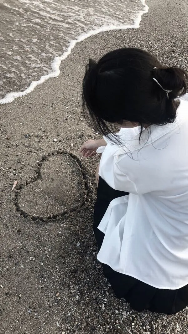 Gái xinh quay lưng vẽ trái tim trên cát vàng bằng ngón tay áo trắng dài buông xõa tóc đen nhẹ trong gió biển tạo dáng dịu dàng.