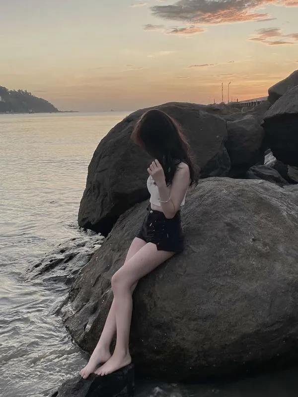 Người đẹp giấu mặt bên bờ cát đứng nghiêng trên tảng đá lớn mặc áo croptop trắng thả dáng tự do.
