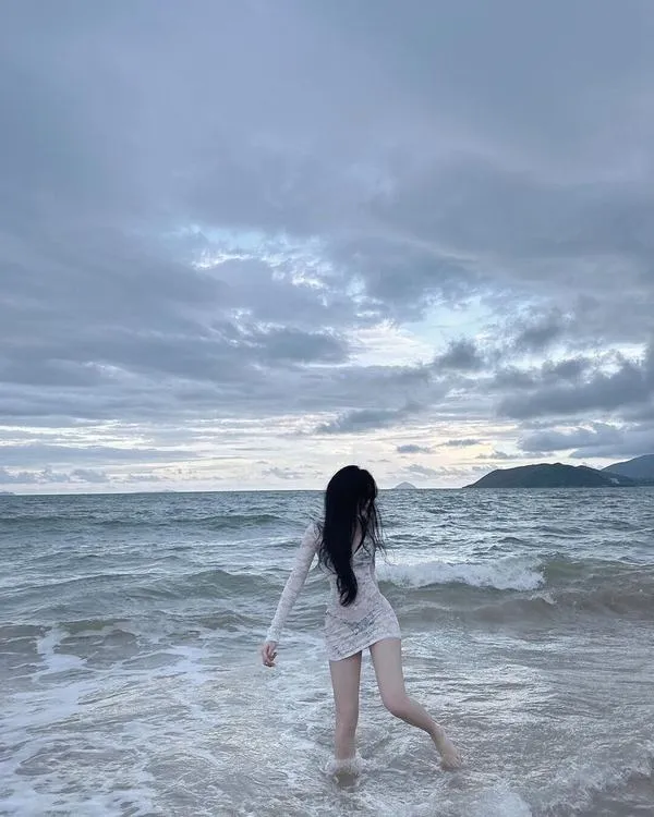 Ảnh gái ngồi xổm trên bãi đá mặc bikini trắng tay nâng tóc dài nhìn ra biển xanh ngắt.