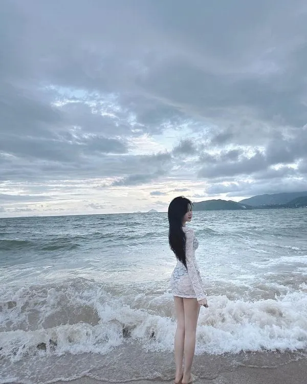 Gái xinh đứng quay lưng bên sóng mặc áo hai dây trắng tay che tóc sau gáy tạo dáng mượt mà.