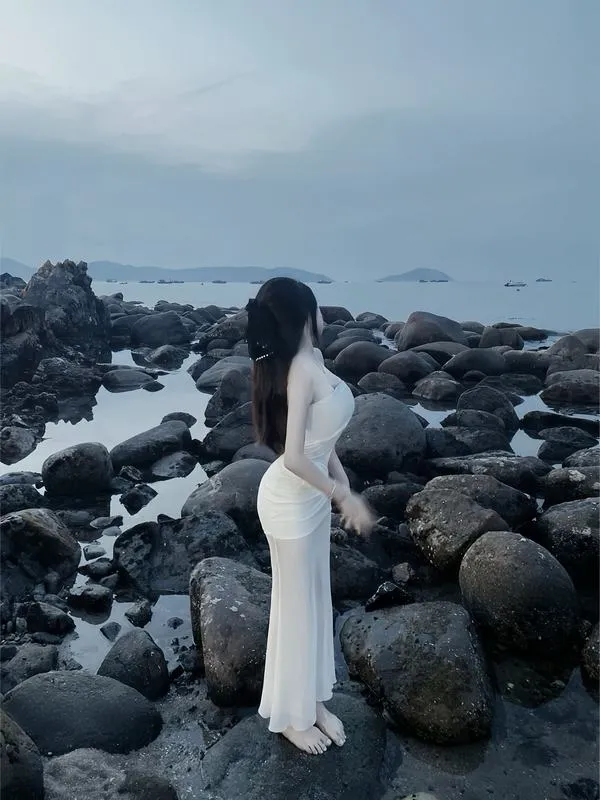 Quý cô bí ẩn ven biển ngồi nghiêng trên ghế dài mặc váy trắng bồng bềnh hoàng hôn dịu dàng.