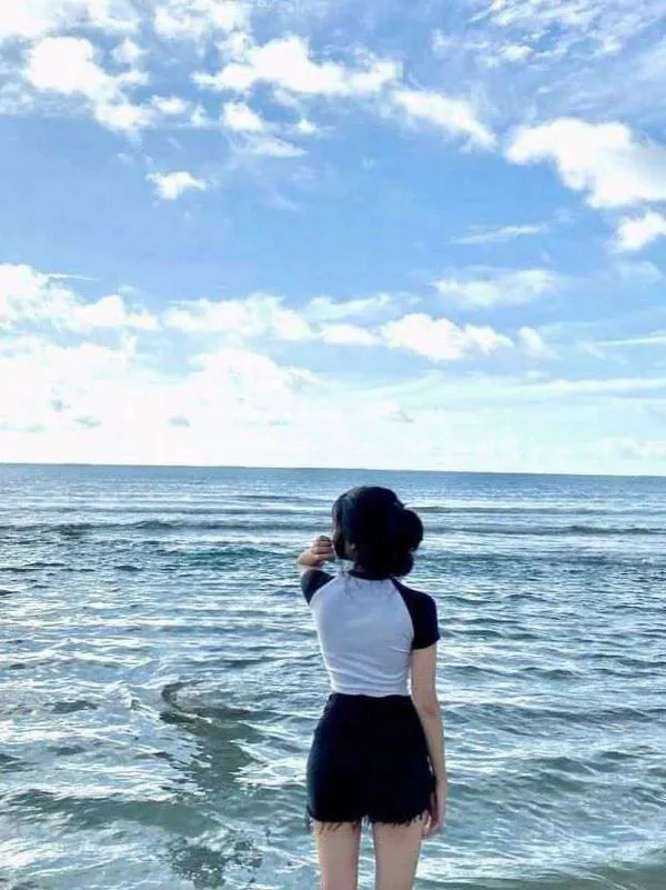 Ảnh gái đứng nghiêng trên bãi đá mặc áo hai dây đen tay vuốt tóc dài hoàng hôn ấm áp.