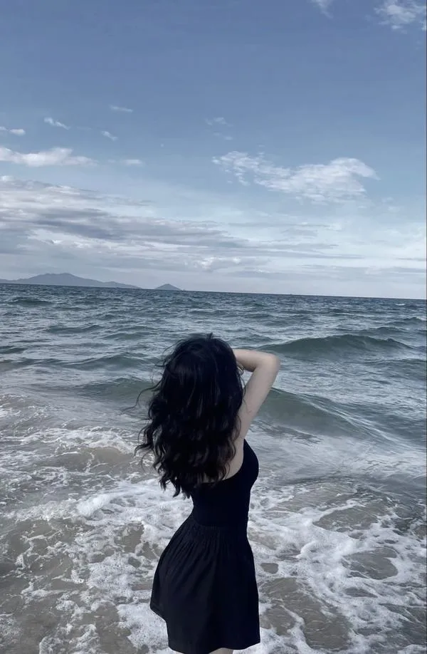 Gái xinh quay lưng nhìn biển mặc váy maxi trắng tay cầm ly nước tóc dài xõa vai.