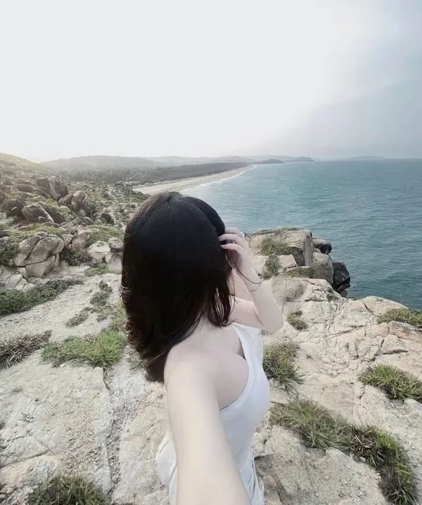 Ảnh gái đứng quay lưng bên sóng mặc áo trắng ngắn tay vuốt tóc dài gió biển mát rượi.