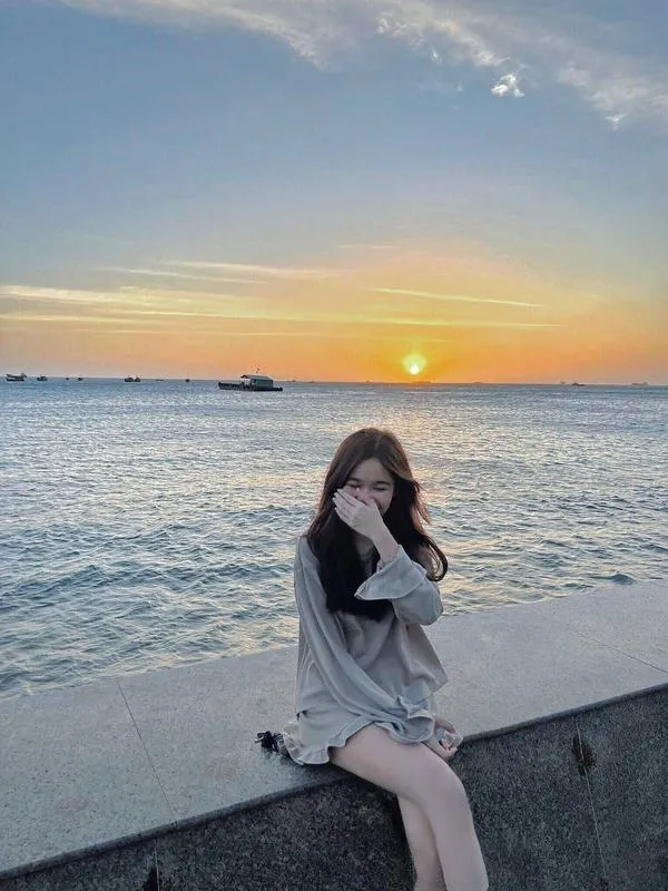 Hot girl che mặt bằng tay ngồi trên ghế dài bãi biển mặc váy trắng bồng bềnh hoàng hôn rực rỡ.