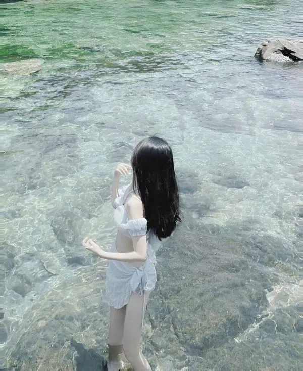 Gái đẹp ngồi xổm bên sóng mặc áo croptop trắng quần jeans ngắn tóc dài che bờ vai trần.