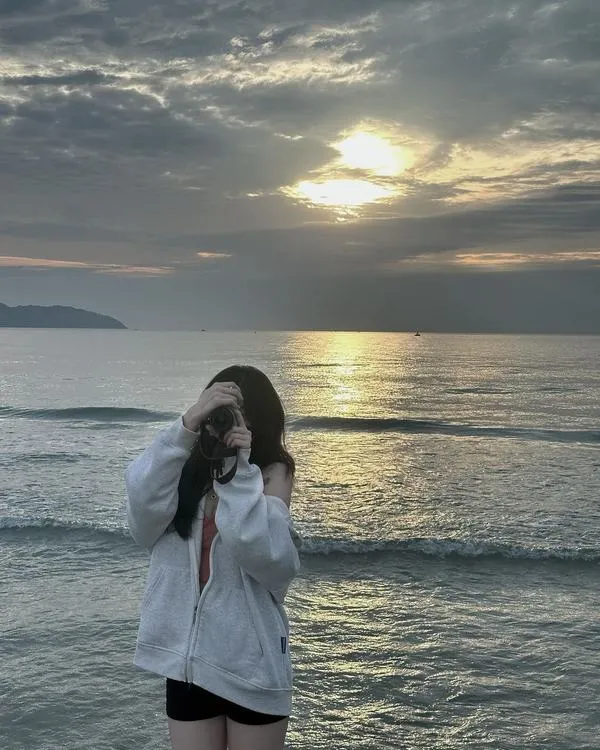 Ảnh gái đứng nghiêng bên rặng đá mặc bikini trắng tay che mặt tóc dài bay gió biển mát.