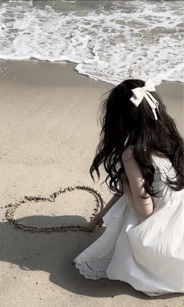 Gái xinh ngồi trên đá ven sóng mặc váy trắng dài tóc xõa vai tay khẽ chạm nước tạo dáng nhẹ nhàng.