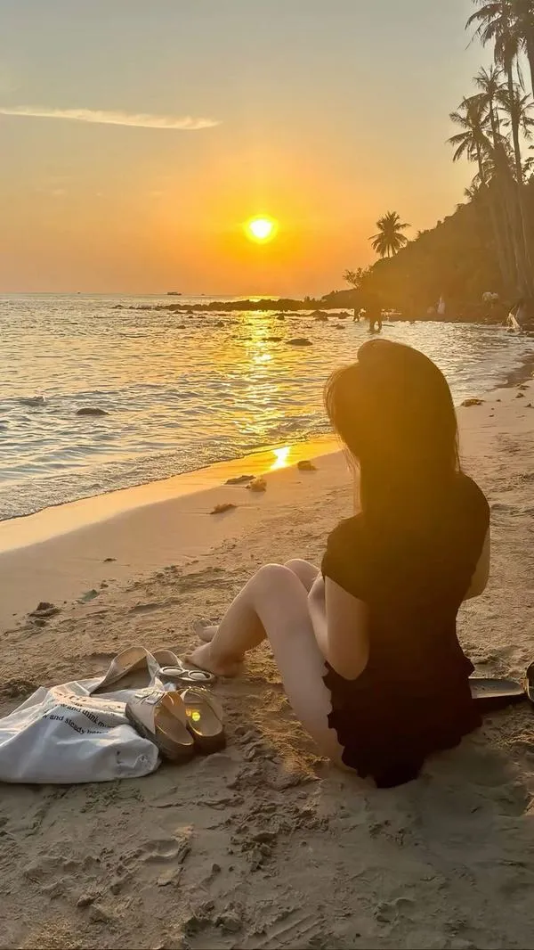 Ảnh gái ngồi nghiêng vẽ trái tim trên cát bằng tay mặc váy trắng dài tóc đen dài dưới nắng chiều.