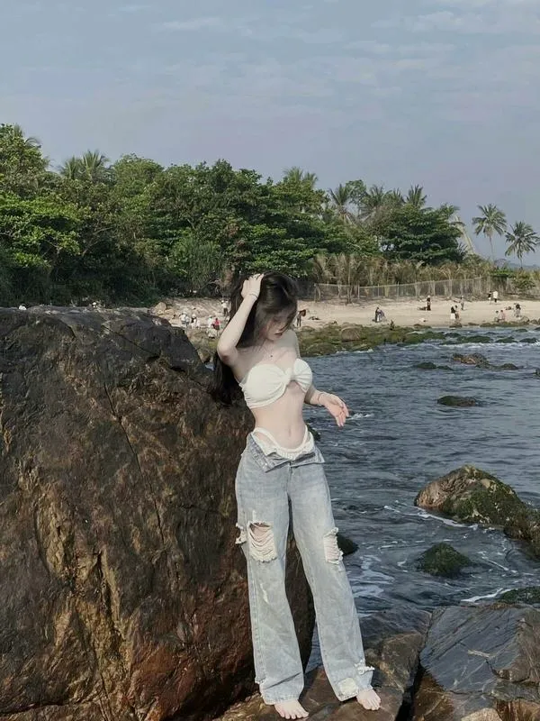 Ảnh gái ngồi quay lưng trên đá ven biển mặc bikini trắng tay cầm điện thoại tóc dài đen xõa vai.