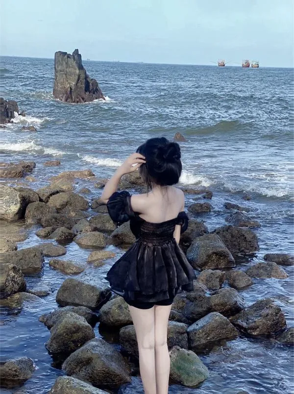 Ảnh gái ngồi nghiêng trên cát vàng hoàng hôn mặc bikini đen một mảnh tay chống đất nhìn ra biển xa.