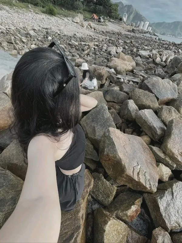 Người đẹp giấu mặt bên bờ cát thả dáng ngồi co gối trên đá biển mặc váy đen ngắn bờ vai trần gió thổi nhẹ.