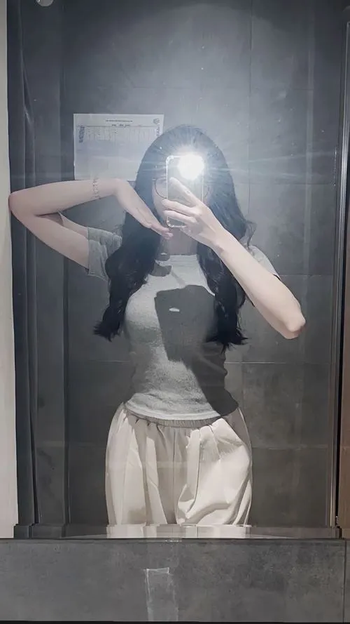 Thiếu nữ ẩn danh mặc áo crop trắng khoe eo hai tay nâng điện thoại che mặt uốn người trước gương.