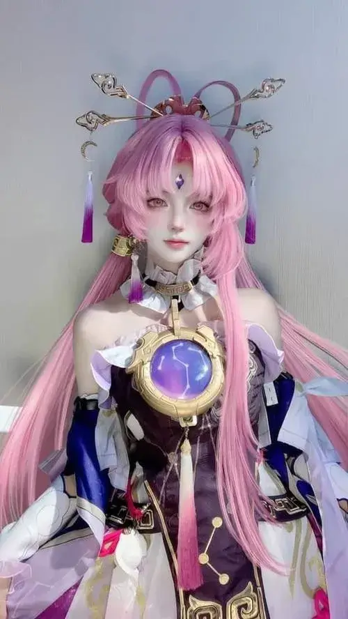 Ảnh thiếu nữ cosplay game Liên Quân với tóc hồng dài, đạo cụ quả cầu trước ngực và ánh nhìn dịu trong không gian trong nhà.