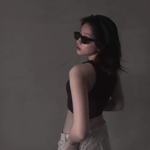 Gái xinh tóc đen dài đứng nghiêng người đeo kính đen gọng vuông nhìn xa xăm trong ánh sáng mờ ảo với vai trần nhẹ.