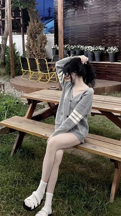 Gái xinh ngồi nghiêng trên ghế gỗ picnic ngoài trời mặc áo xám tay dài tóc búi cao một bên tay che trán nhìn xa xăm trong nắng dịu.