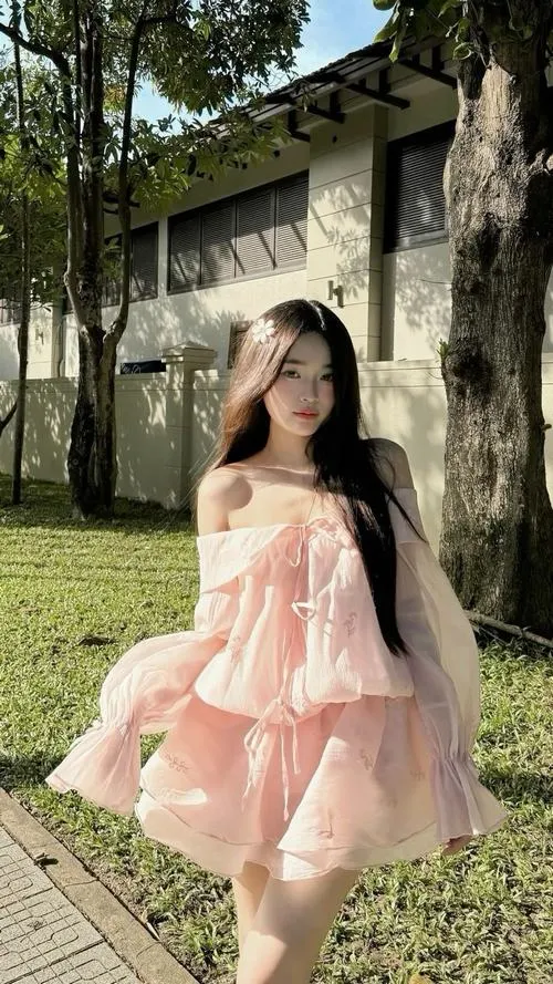 Ảnh gái ngồi trên cỏ xanh mặc váy hồng trễ vai buông vai nhẹ mái tóc đen dài xõa tự nhiên ánh mắt hướng sang bên trong vườn ngoài trời xanh mát rạng rỡ.