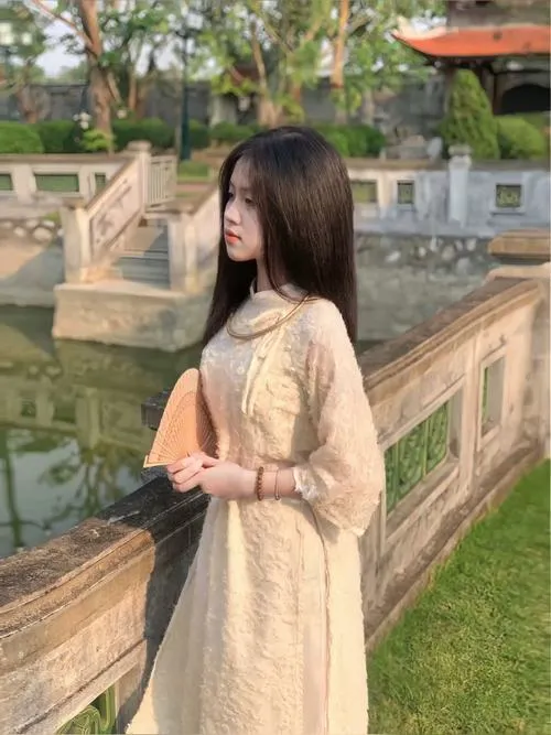 Gái xinh đứng nghiêng bên lan can đá mặc áo dài kem nhạt ôm dáng mảnh mai tóc dài buông xõa nhẹ nhàng dưới nắng xuân dịu dàng trong thiếu nữ diện đồ tết.