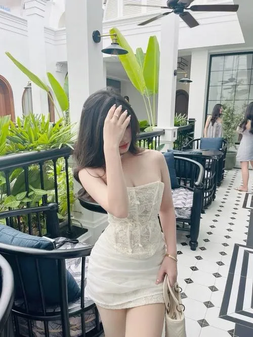 Gái xinh ngồi nghiêng trên ghế sofa trắng trong phòng khách sang trọng mặc váy trắng ôm sát vai trần tóc dài xõa vai tay cầm túi xách nhìn nhẹ sang bên toát lên vẻ dịu dàng nữ tính mỹ nhân mặc váy.