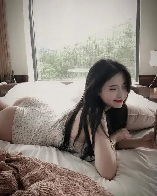 Gái xinh nằm nghiêng trên giường trắng với tay chống cằm mái tóc đen dài xõa tung mặc váy ngủ satin trắng mỏng nhẹ ánh mắt hướng camera nụ cười dịu dàng.