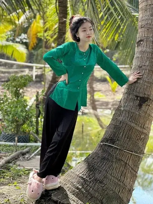 Ảnh gái mặc áo xanh lá đứng tựa cây cọ, tay chống hông nhìn ngang, tóc búi cao gọn gàng giữa nắng vàng rực.