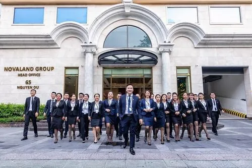 Ảnh chuyên viên địa ốc Novaland xếp hàng trước tòa nhà lớn với logo thương hiệu nổi bật, dáng đứng ngay ngắn và nụ cười thân thiện rất cuốn hút