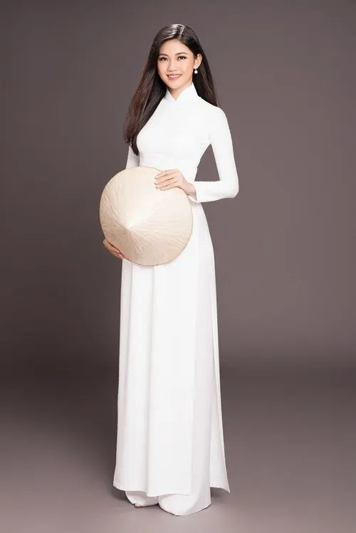 Ảnh gái mặc áo dài trắng đứng nghiêng nhẹ cầm nón lá che một bên má, mái tóc dài xõa vai nhẹ nhàng giữa ánh nắng vàng.