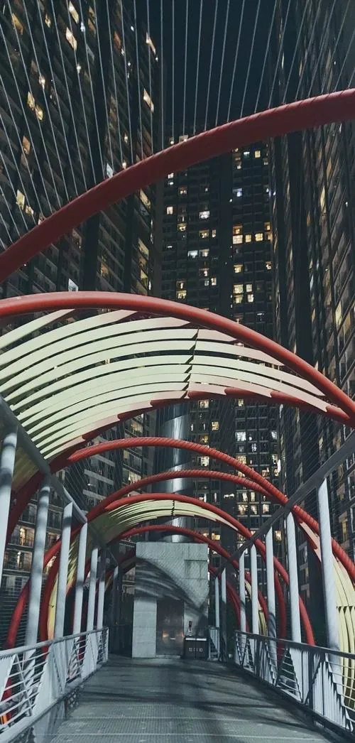 ảnh công viên ven sông lối đi mái vòm đỏ uốn cong, người đi bộ thảnh thơi dưới bóng cây, ánh sáng dịu phủ lên khung cảnh xanh mát ngay công viên Vinhomes Central Park sang trọng