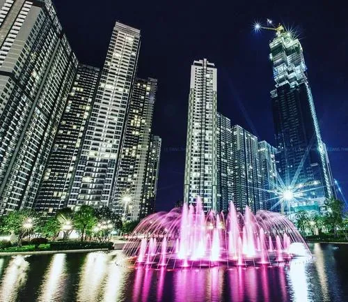 Một góc chụp ban đêm với các tòa nhà sáng đèn, phía trước là quảng trường và đài phun nước phản chiếu ánh sáng, tạo khung cảnh lung linh tại công viên ven sông Vinhomes