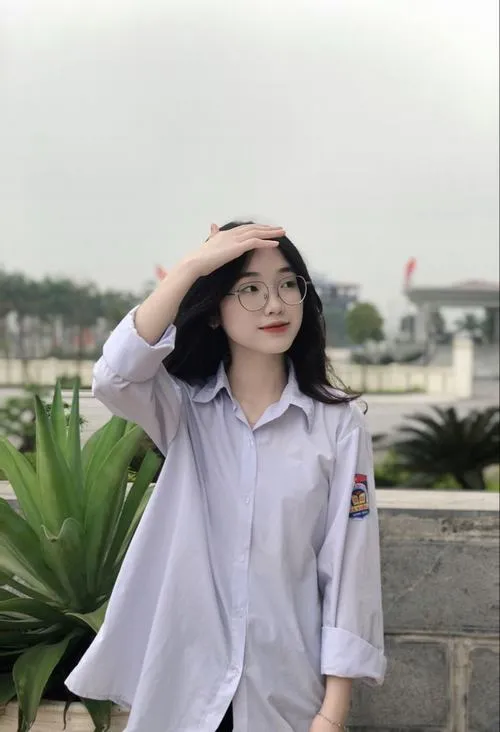 Gái xinh tóc dài mặc áo sơ mi trắng đứng ngoài sân trường ngày nắng, tay che nhẹ trán nhìn camera với nụ cười rạng rỡ toát lên vẻ tươi mới của thiếu nữ sinh năm 2011.
