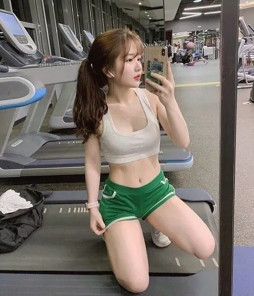 Gái xinh đứng tự sướng gương phòng gym mặc crop top trắng quần short xanh lá, vẻ mặt tươi tỉnh đầy năng lượng quyến rũ.