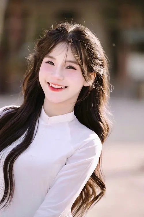 Gái xinh tóc dài nâu óng mặc áo trắng cổ cao mỉm cười rạng rỡ nhìn thẳng vào ống kính trong ánh sáng dịu.