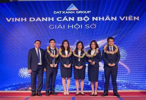 Ảnh nhân sự nổi bật trong bộ đồng phục xanh đồng bộ đứng thành hàng trên sân khấu lớn, nở nụ cười tươi trước backdrop thương hiệu tạo nên khung hình chuyên nghiệp và rạng rỡ của tập đoàn đất xanh