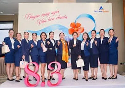 Nhóm nhân viên nữ tươi tắn tạo dáng bên cụm chữ trang trí trong sảnh sự kiện, váy công sở và blazer xanh giúp tổng thể thêm thanh lịch và bắt mắt