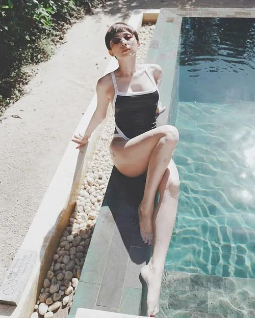 Gái xinh mặc bikini đen cá tính đứng trên ván lướt sóng, nghiêng người khoe vóc dáng săn chắc và bờ vai trần.