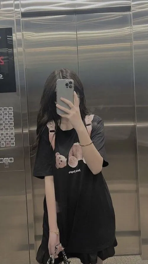 Hot girl tóc nhuộm màu cà phê mặc váy tennis trắng đứng trước backdrop trắng cầm điện thoại.