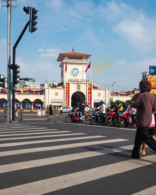 ảnh địa danh lịch sử Sài Gòn ghi lại khoảnh khắc người dân băng qua đường trước cổng, vài chiếc xe máy dừng lại, tạo nên nhịp sống quen thuộc của khu chợ sầm uất