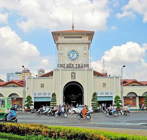 Một góc nghiêng khác của cổng chợ, xe máy chạy ngang liên tục, ánh sáng chiều chiếu vào làm nổi bật lớp sơn vàng đã nhuốm màu thời gian