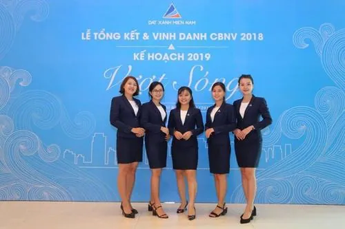 Bốn nữ nhân viên đứng thành hàng trước backdrop xanh thương hiệu, chân váy công sở ôm dáng cùng nụ cười tươi làm nổi bật nét đẹp thanh lịch của dàn cực phẩm Đất Xanh