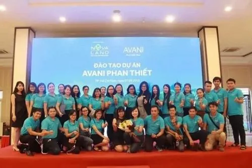 Ảnh chuyên viên tư vấn chụp tập thể tại hội trường lớn với ánh đèn vàng ấm, đồng phục xanh trắng tạo tổng thể tươi sáng và nổi bật