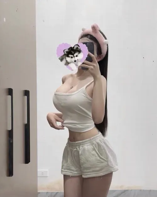 Cô nàng tóc dài đen óng ả đang tựa tay lên cằm trong phòng ngủ sáng, mặc áo hai dây xám ôm sát tôn đường cong, ánh mắt hướng thẳng camera với nụ cười nhẹ nhàng thu hút.