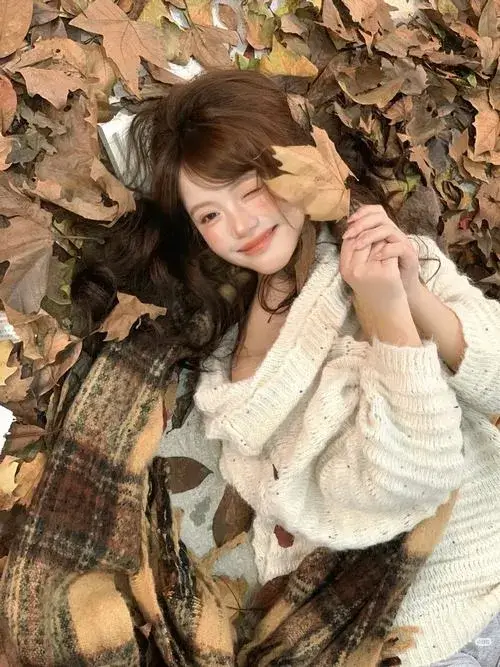 Gái xinh tóc nâu buông xõa nhẹ nhàng ôm mặt mỉm cười rạng rỡ trong áo len trắng giữa nền lá cây xanh tươi ấm áp.